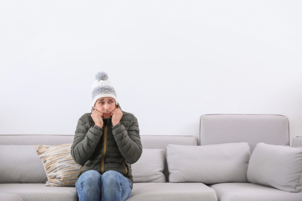 Young man wearing warm clothes freezing on sofa at home depicting furnace turns off after reset