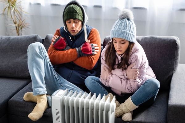 couple feeling cold wearing full winter clothes indoor sitting in front of a space heater due to malfunctioning furnace